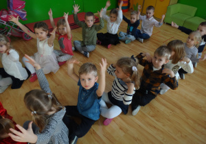 Przedszkolaki siedzą na podłodze unosząc ręce w górę